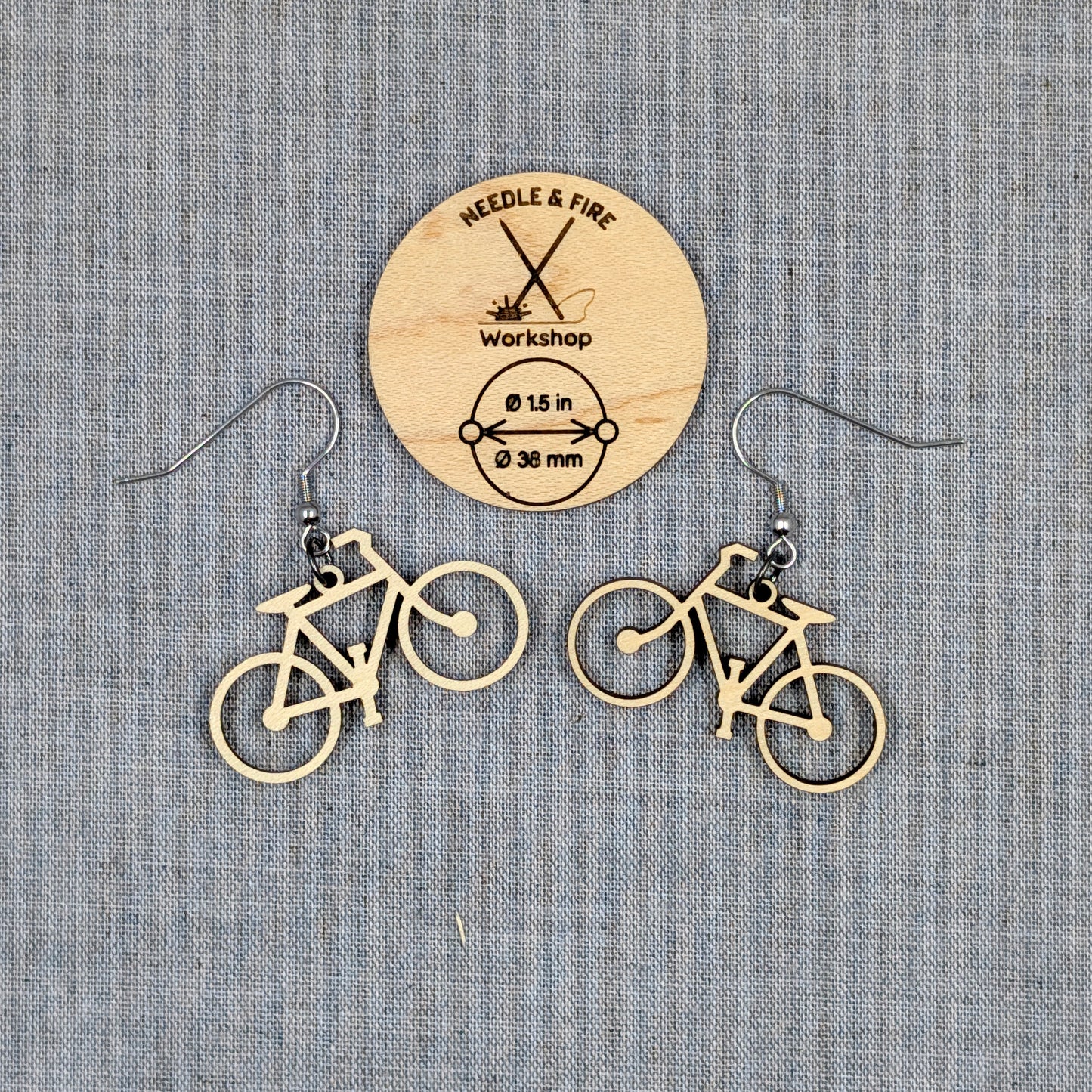A pair of bicycle-shaped dangle earrings made of light natural wood, displayed on a textured gray fabric background with a circular wooden brand card for Needle & Fire Workshop.