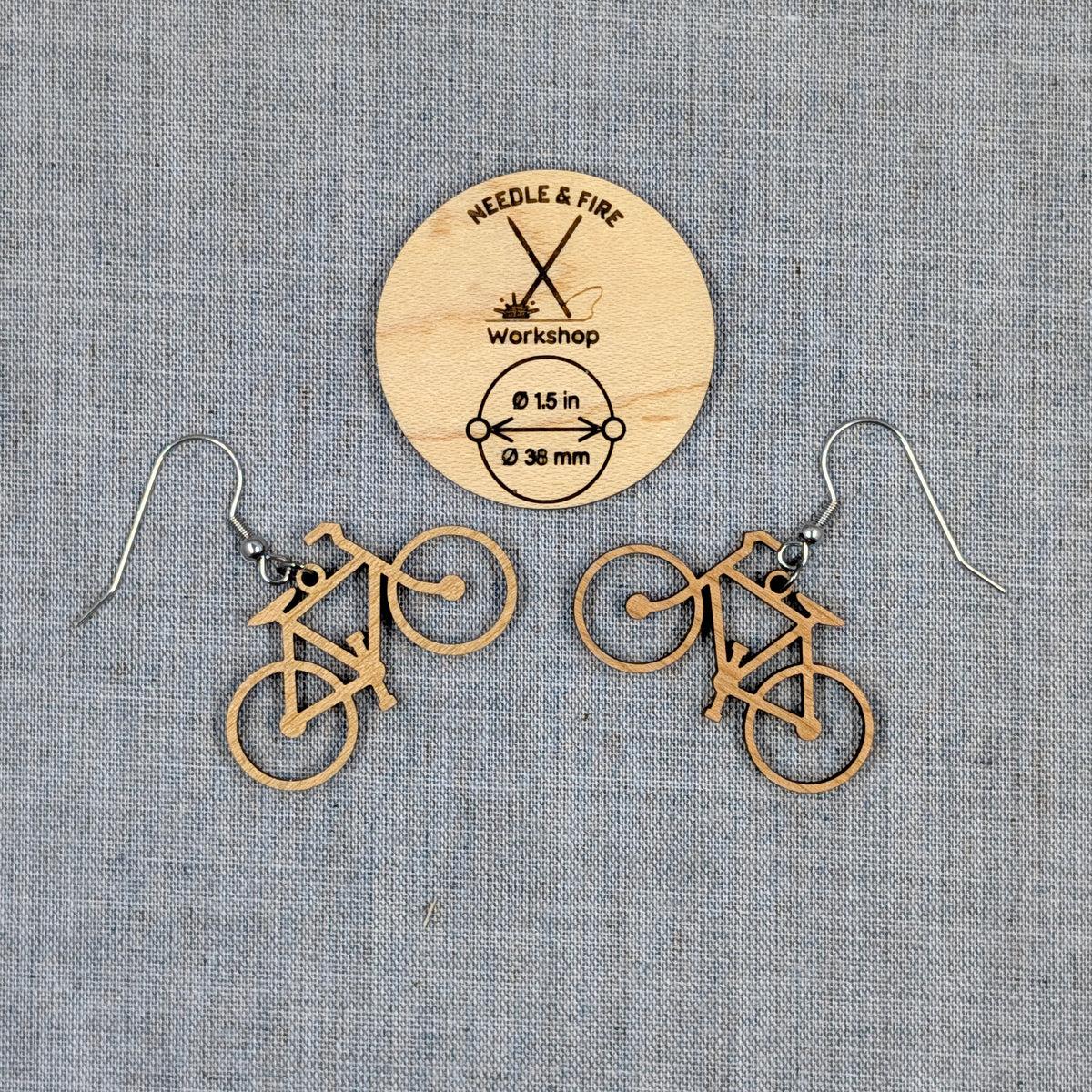 A pair of bicycle-shaped dangle earrings made of natural wood, displayed on a textured gray fabric background with a wooden brand tag reading Needle & Fire Workshop.