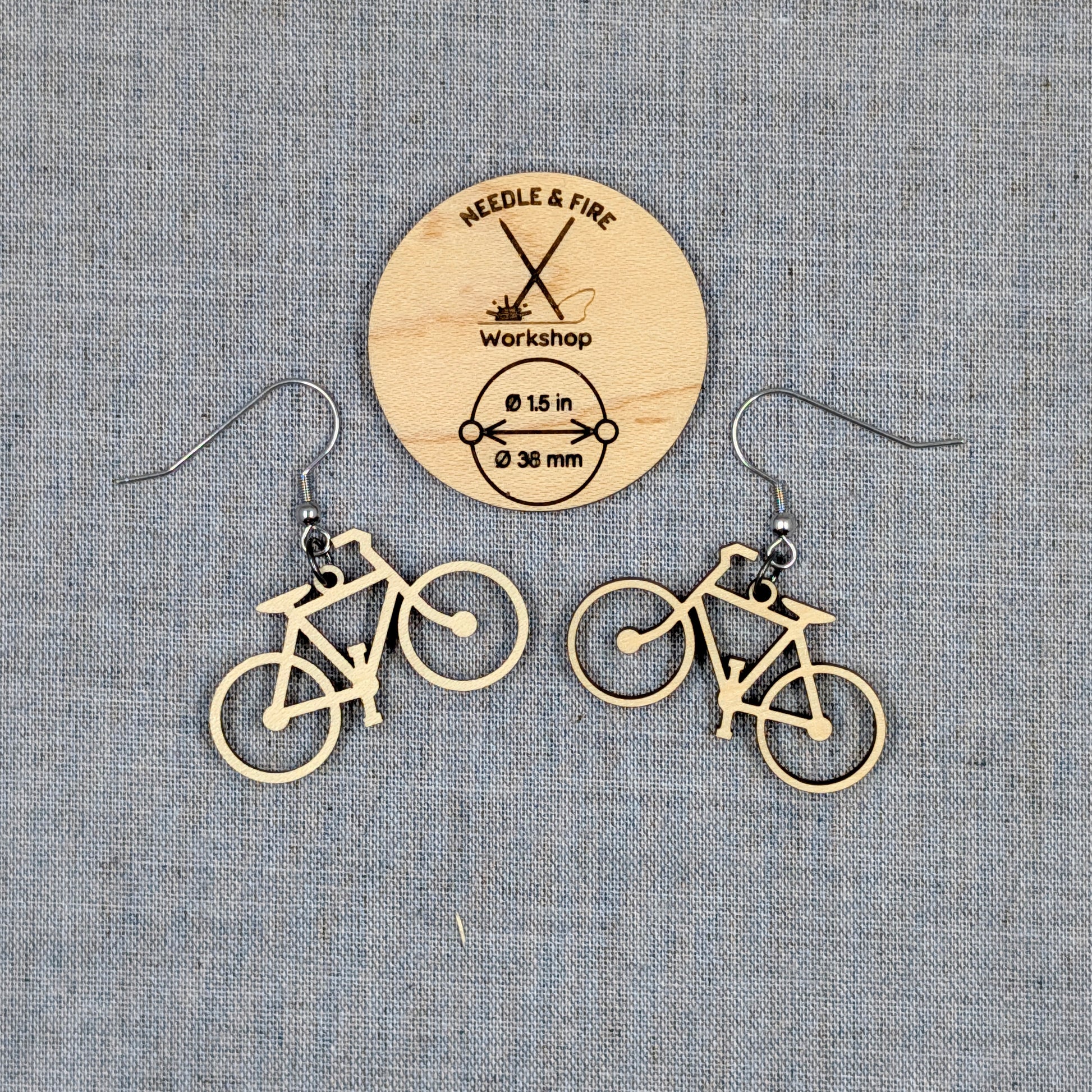 A pair of bicycle-shaped dangle earrings made of light natural wood, displayed on a textured gray fabric background with a circular wooden brand card for Needle & Fire Workshop.