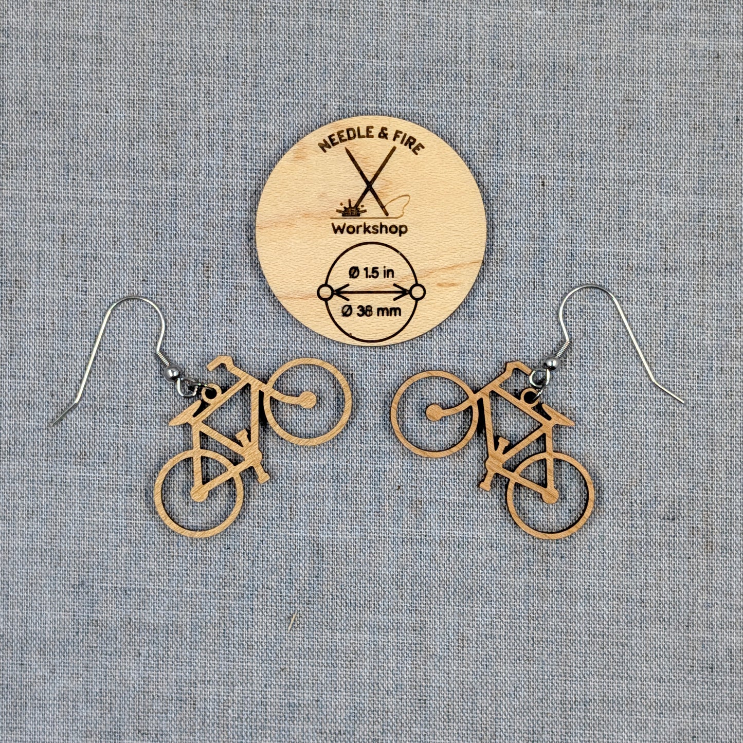 A pair of bicycle-shaped dangle earrings made of natural wood, displayed on a textured gray fabric background with a wooden brand tag reading Needle & Fire Workshop.