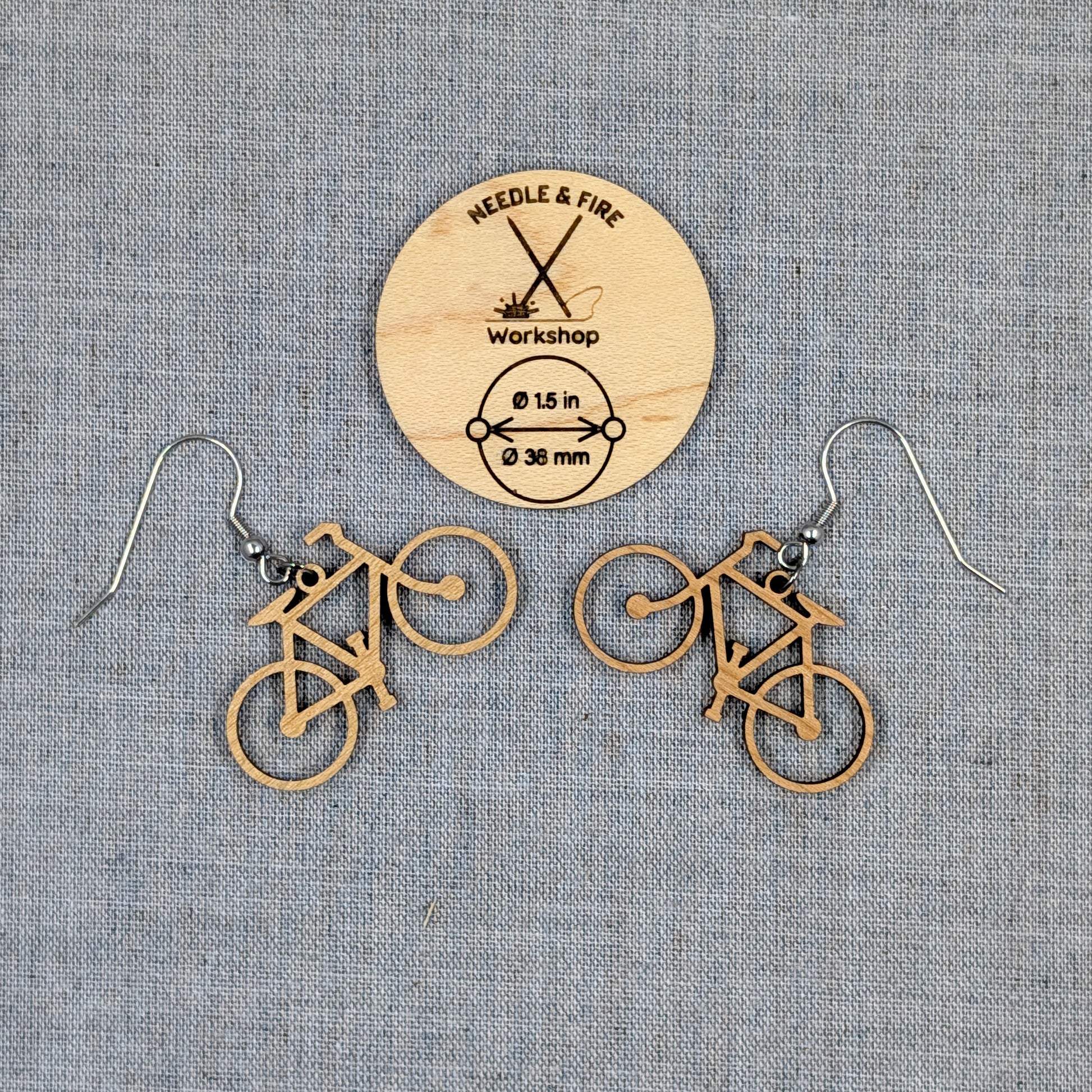 A pair of bicycle-shaped dangle earrings made of natural wood, displayed on a textured gray fabric background with a wooden brand tag reading Needle & Fire Workshop.