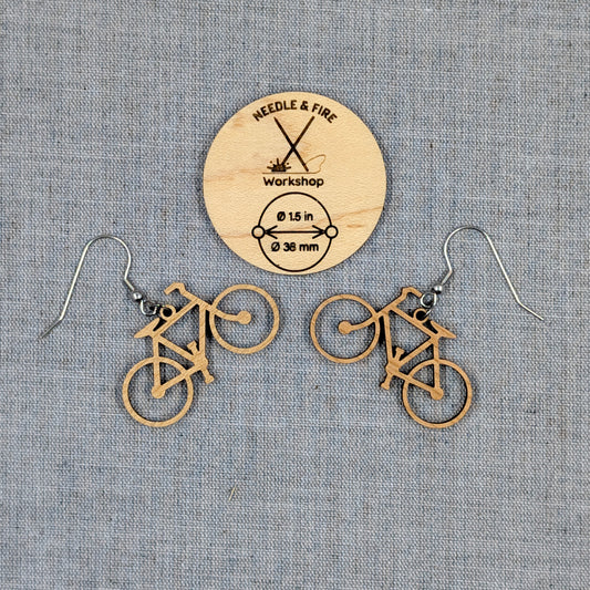 A pair of bicycle-shaped dangle earrings made of natural wood, displayed on a textured gray fabric background with a wooden brand tag reading Needle & Fire Workshop.